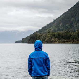A person in a blue jacket stands by a misty New Zealand lake, surrounded by mountains.